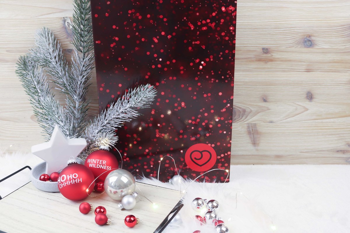 Dessous Adventskalender von Lovehoney 2019: geschlossene dunkelrote Box mit roten Lichtpunkten und Herz-Logo, davor Weihnachtskugeln, Stern und Tannenzweig.